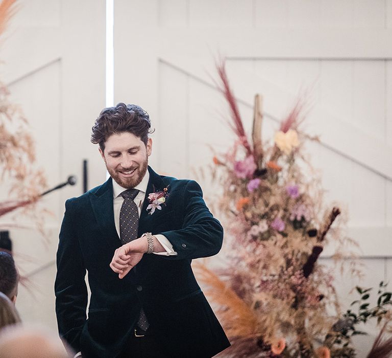 Groom in velvet suit checks his watch for the time as he stands in front of colourful dried flower columns on wedding day 