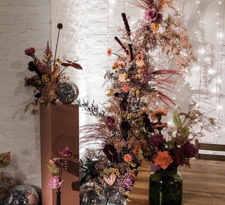 Purple and orange flowers in vases and in a flower column with disco balls and fairy light back drop for wedding reception decor |