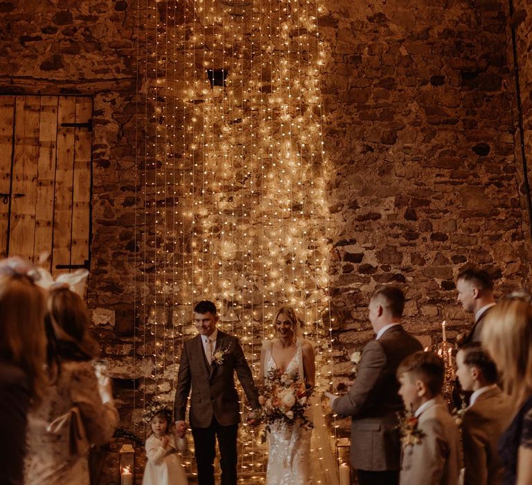 Bride and groom stand in front of fairy light backdrop at Eden Barn with the groom holding hands with their daughter in white dress and flower crown