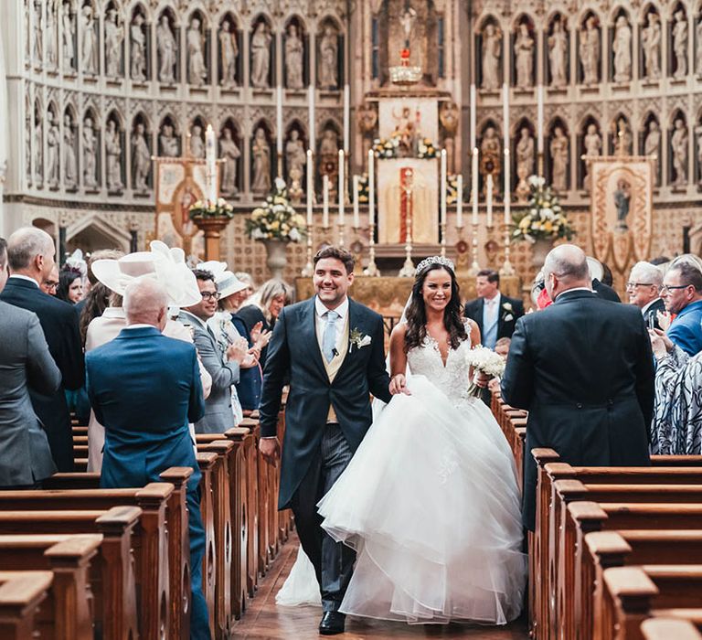 Bride in princess wedding dress with Princess Diana style crown walks back down the aisle with the groom in a morning suit with pale blue tie