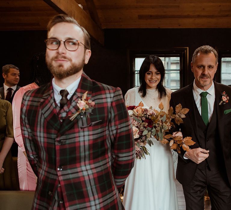 Groom wearing bespoke tartan suit awaits his bride at the end of the aisle as she walks arm in arm with her father and holds floral bouquet