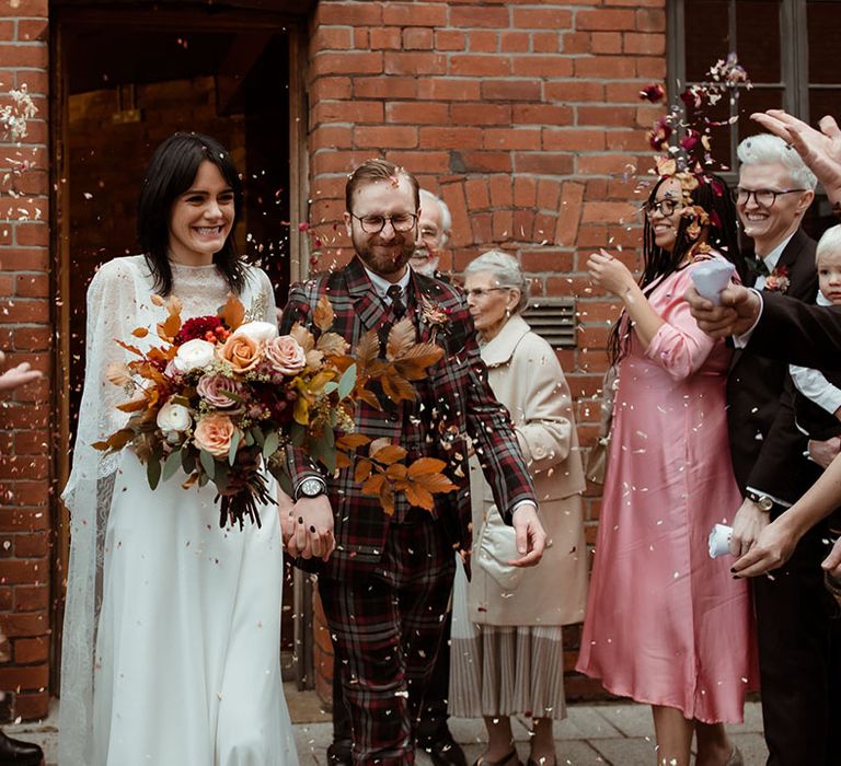 Bride & groom leave The Chimney House as wedding guests throw confetti around them and bride holds Autumnal floral bouquet 