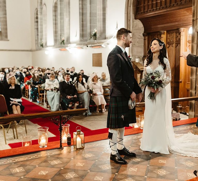 Bride wears long sleeved lace gown and groom wears tartan kilt as they stand at the altar in church during wedding ceremony 