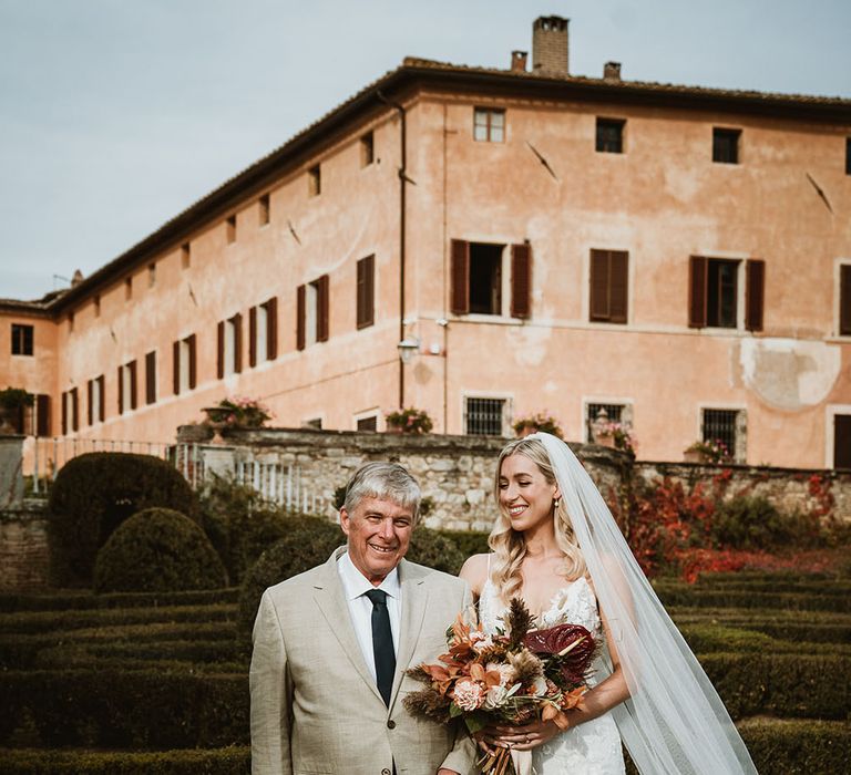 Bride holding tropical and colourful bouquet stands beside her father before walking down the aisle outdoors 