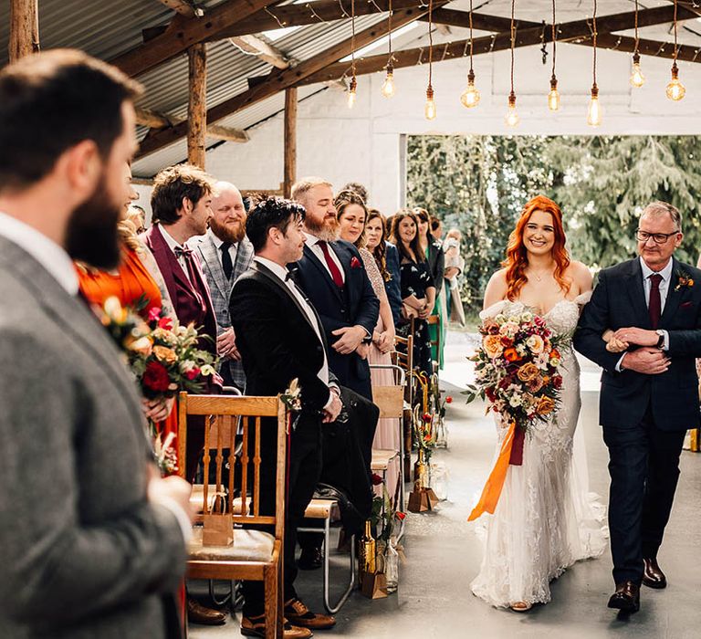 Bride with large red and orange autumnal bouquet is walked down the aisle by her father to meet the groom 