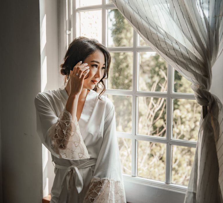 South East Asian bride wears white lace short dressing gown and wears her dark hair pulled back into low bun complete with curled loose tendrils framing her face 