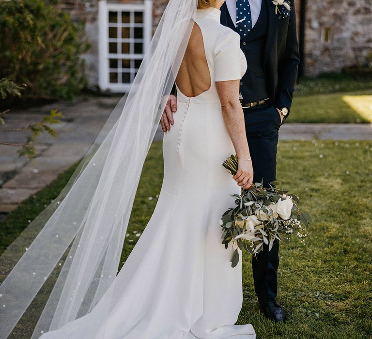 Bride in high neck wedding dress with backless detail and pearl wedding veil with groom in a blue suit 