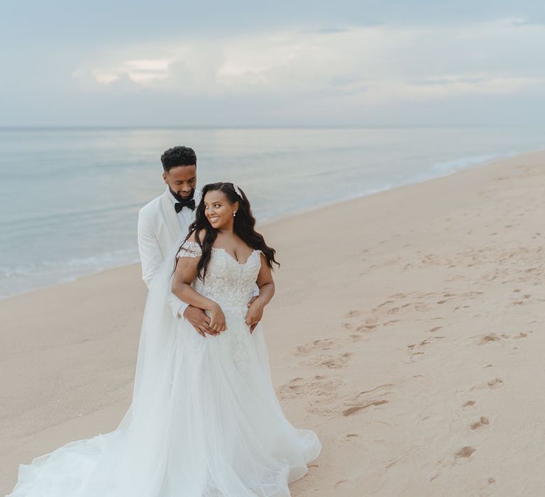 Bride wears off-the-shoulder lace wedding dress as she stands with her groom on the seafront in Phuket 