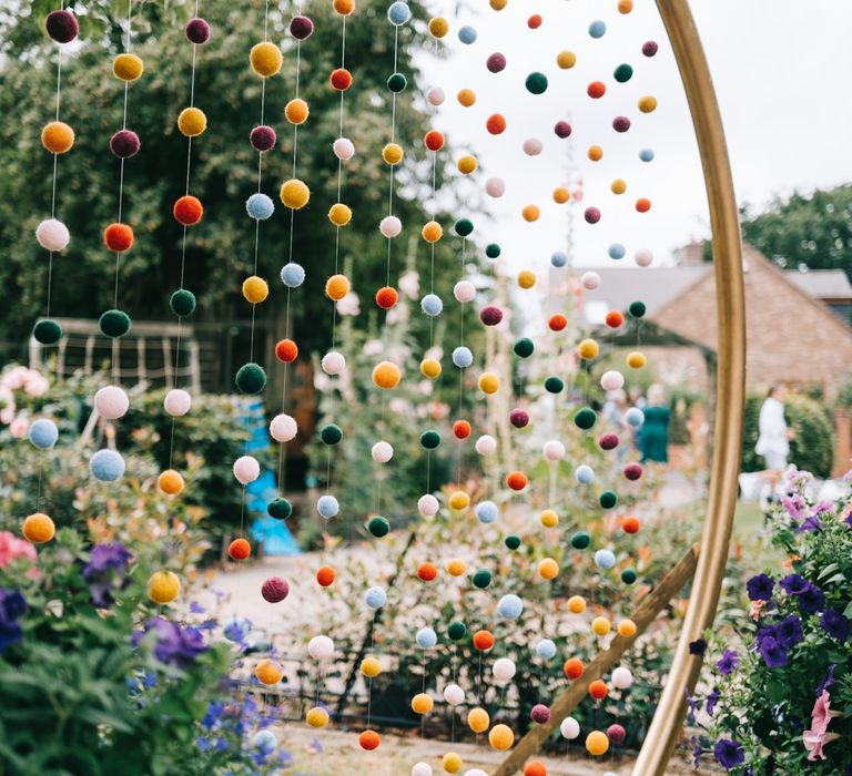 Colourful pom pom wedding decorations hang from gold circular arch 