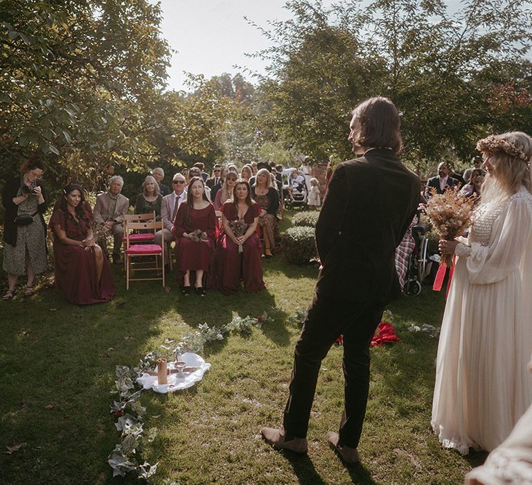 Bride & groom stand surrounded by florals during outdoor wedding ceremony as guests watch on