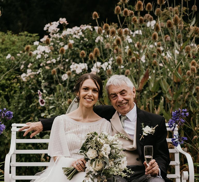 Bride in batwing sleeve lace detail wedding dress and neutral toned bridal bouquet sitting with her father in three piece suit with white boutonniere