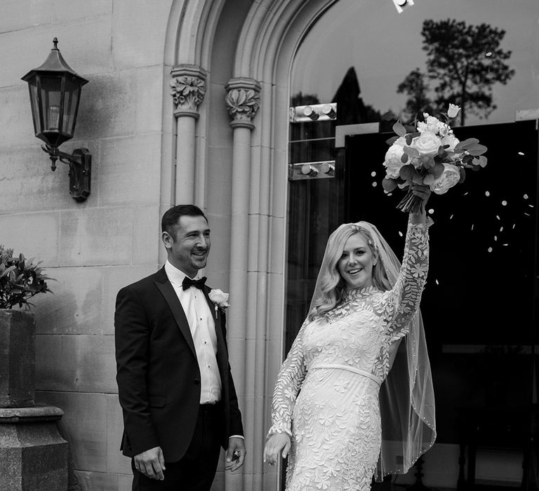 Bride holds up floral bouquet in celebration and stands with her groom in black-tie in black & white image