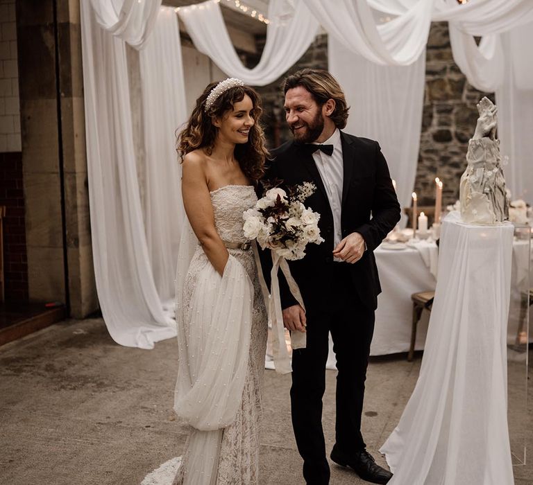 Bride in sleeveless lace wedding dress with pearl details, pearl headband and silver belt standing with groom in black tux by ceiling drapes decoration