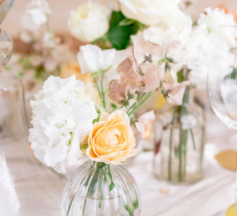 White, yellow and cream flowers in small glass vases decorate the outdoor dining table 