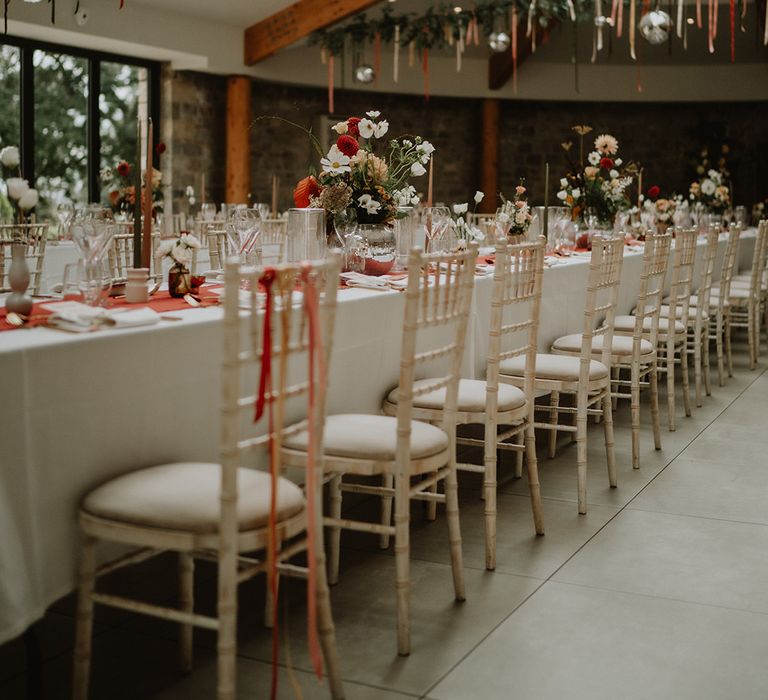 Reception room with streamers, foliage and disco ball decor hanging above white wedding tablescape with pops of 70's-inspired burnt orange and brick red 