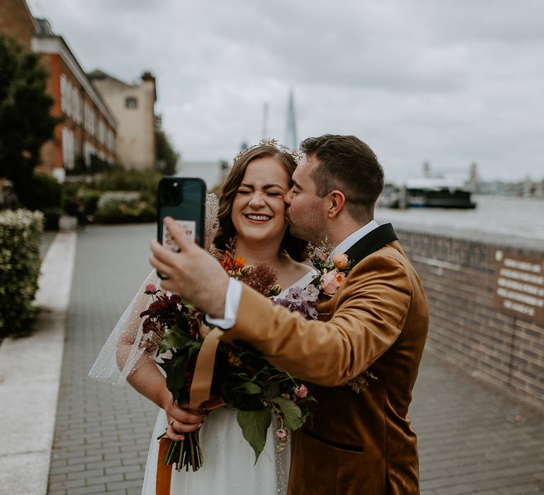 Groom kissed Bride on cheek whilst taking a selfie with Burnt orange themed bouquet and suit