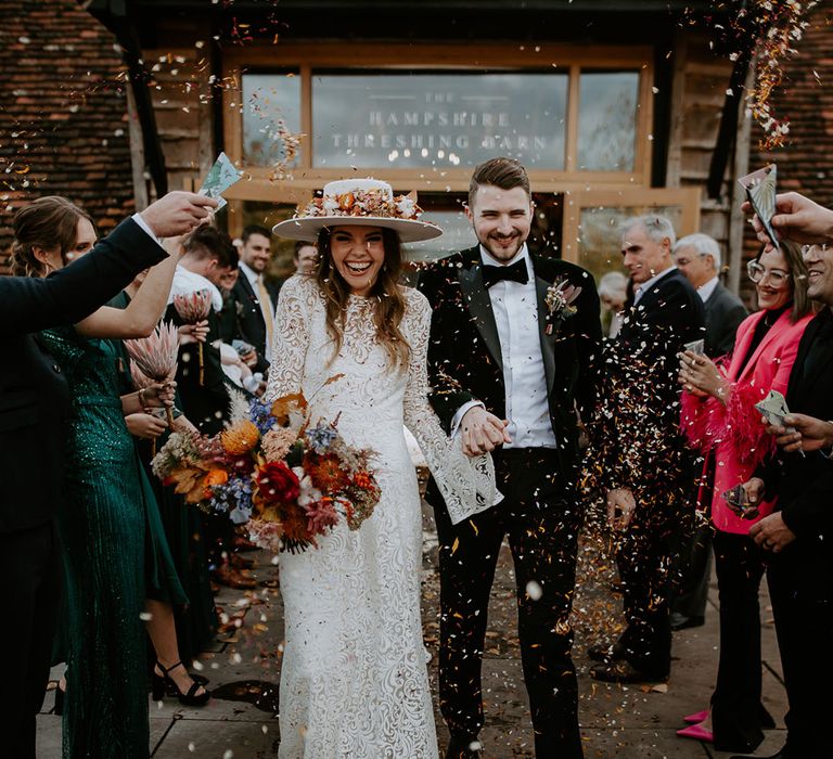 Bride in laser cut lace wedding dress with a hat and colourful bouquet walking with the groom in a velvet tux under confetti 