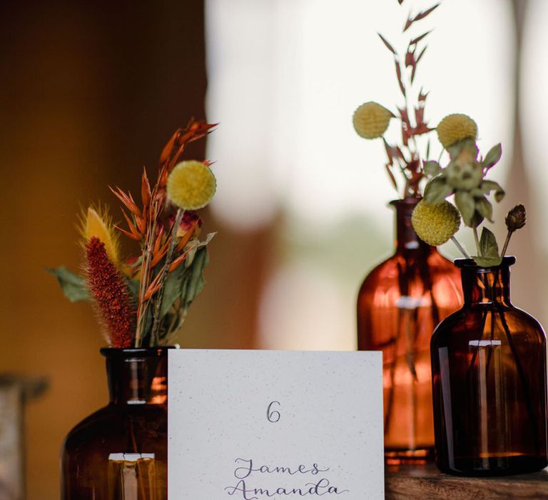 Minimalistic sustainable wedding table number with midnight blue calligraphy resting against dark orange small bottle with dried yellow craspedias, terra cotta celosia seeds, burnt orange wildflowers and green foliage 