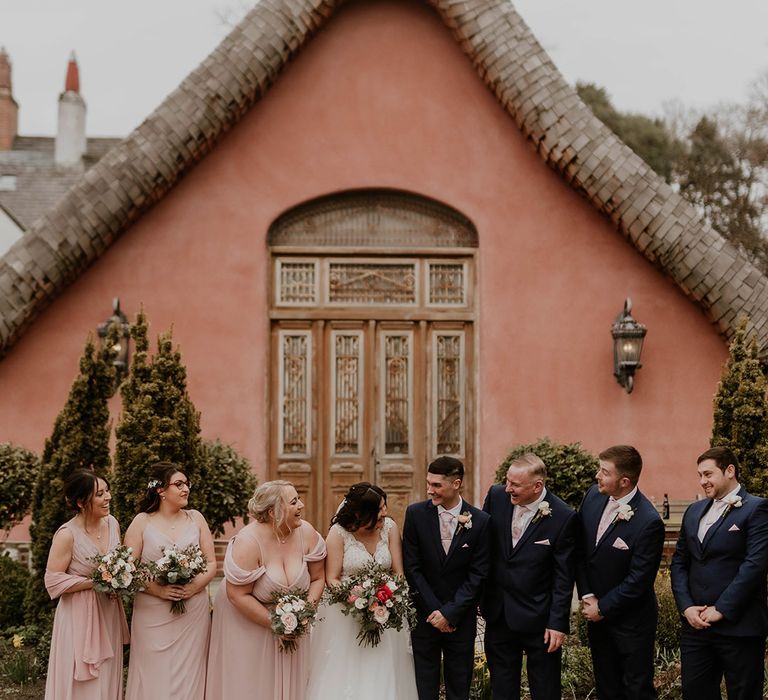 Bridesmaids in pale pink bridesmaid dresses stand with groomsmen in suits and floral buttonholes 