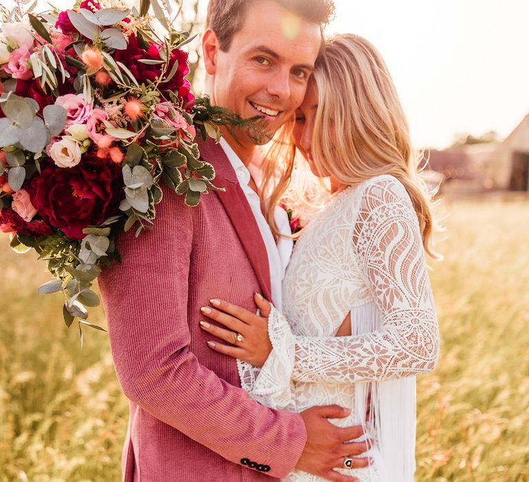 bride in long sleeve lace boho wedding dress with tassels embracing groom in pink corduroy suit