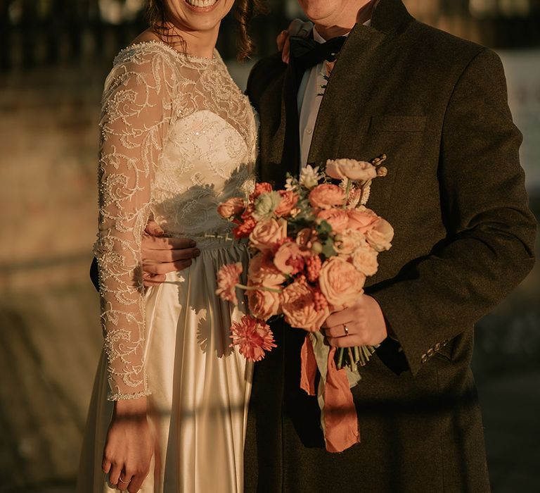 Bride in gold, pearl and emerald bridal crown wearing princess wedding dress with long lace sleeves and groom in black tux holding bouquet with light pink poppies, orange garden roses, eucalyptus and dried flowers