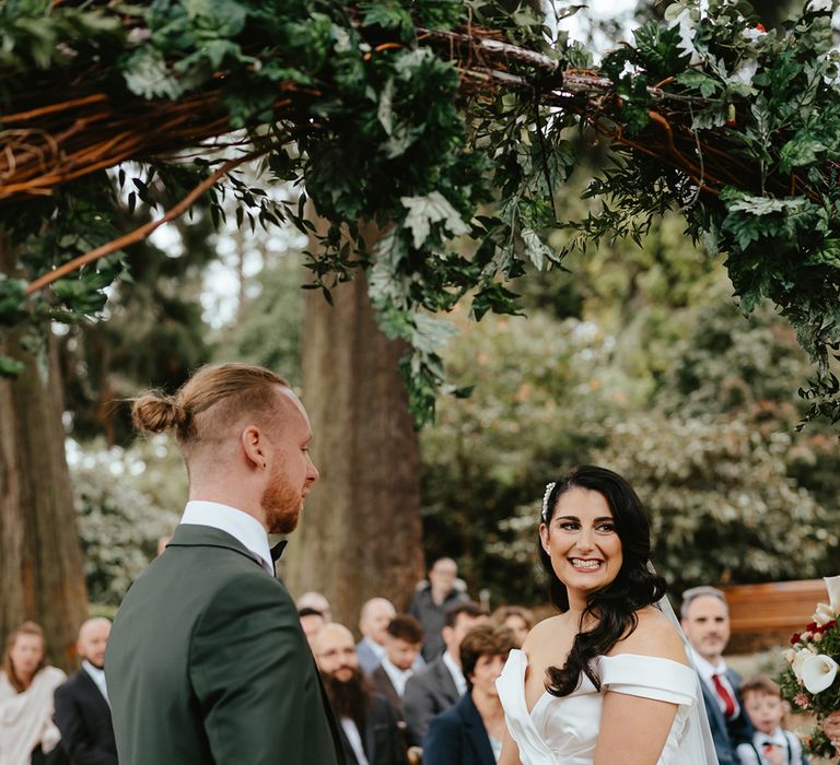 Bride in off-the-shoulder wedding dress and groom in green suit stand beneath foliage archway for outdoor wedding ceremony