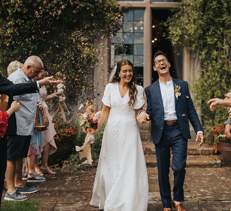 Bride in embroidered wedding dress with short puff sleeves walking with the groom in relaxed navy suit and white shirt as confetti is thrown over them