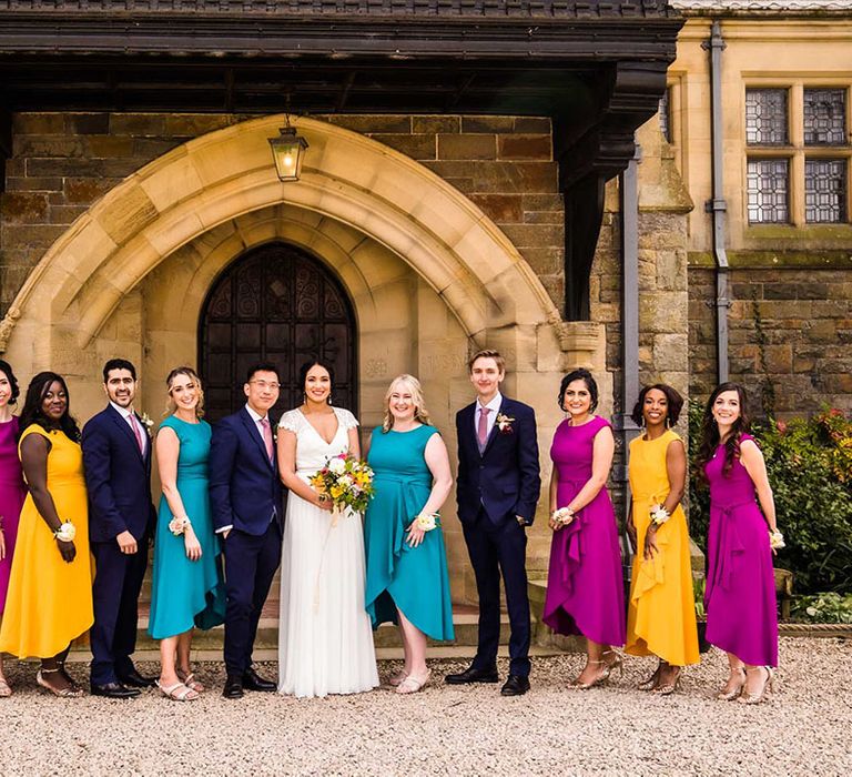 Bride & groom stand with their wedding party of grooms in blue suits beside bridesmaids in yellow, purple and teal mismatched bridesmaid dresses