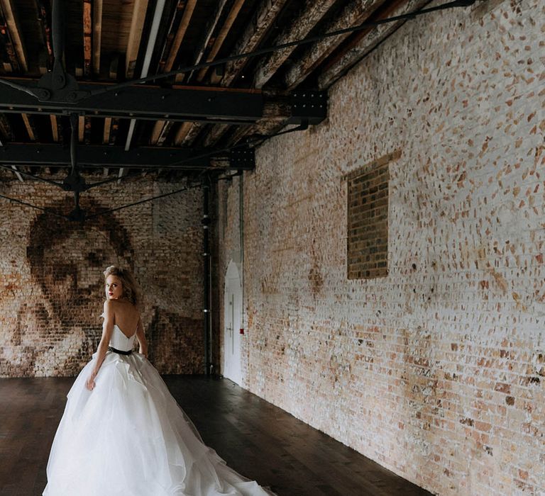 Bride standing in Woolwich Works London wedding venue wearing romantic organza and tulle a-line wedding day bridal ballgown with a flowing skirt, sculpted bandeau top and patterned black belt