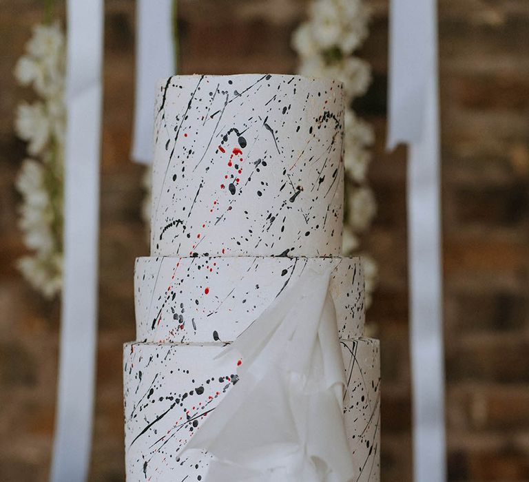 Three tiered buttercream red velvet wedding cake with black paint splatter icing and white layered lace accessory on white cake stand 