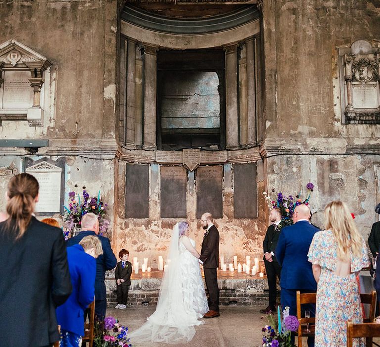 Bride and groom at the alter of London Asylum Chapel holding hands with page boy next to them, surrounded by pillar candles, purple, blue, pink and white flower arrangements with dried flowers and bunny ears