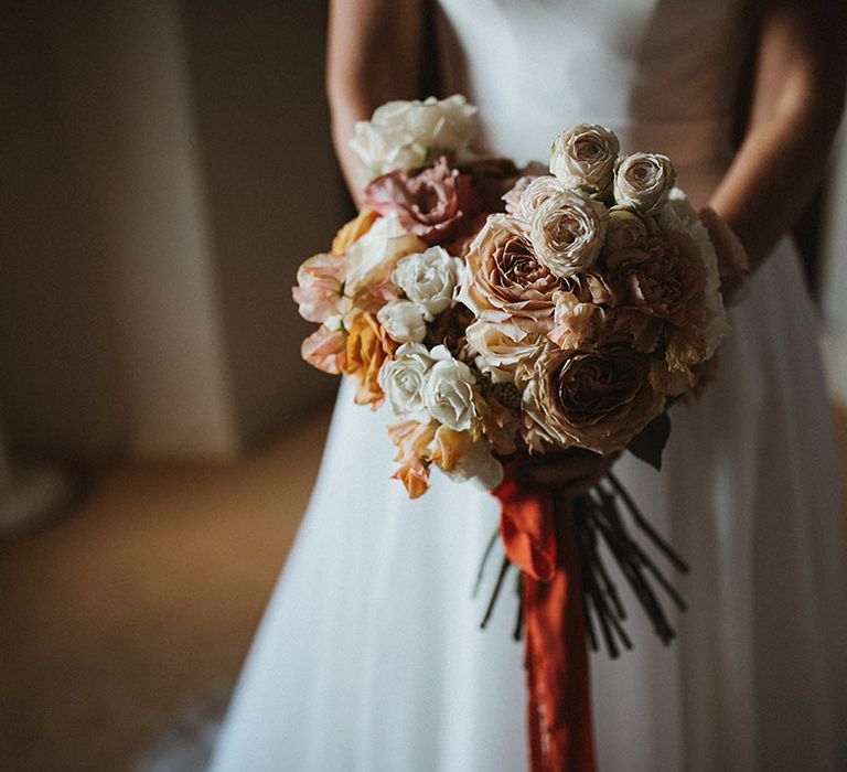 Bride holds pastel rose floral bouquet tied with red silk ribbon 