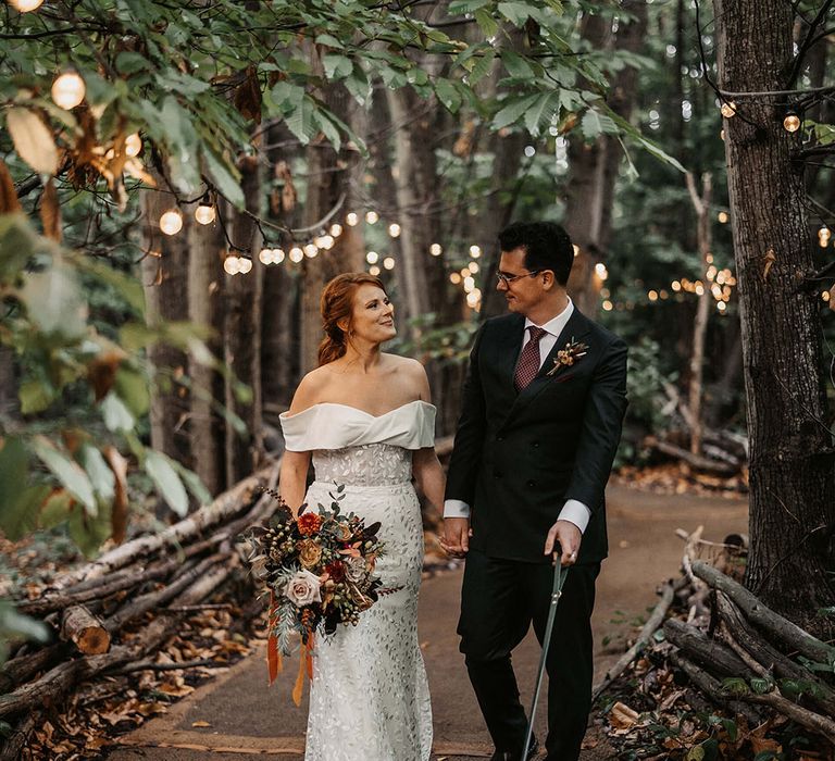 Bride & groom walk through woodland complete with festoon lighting at The Dreys wedding venue