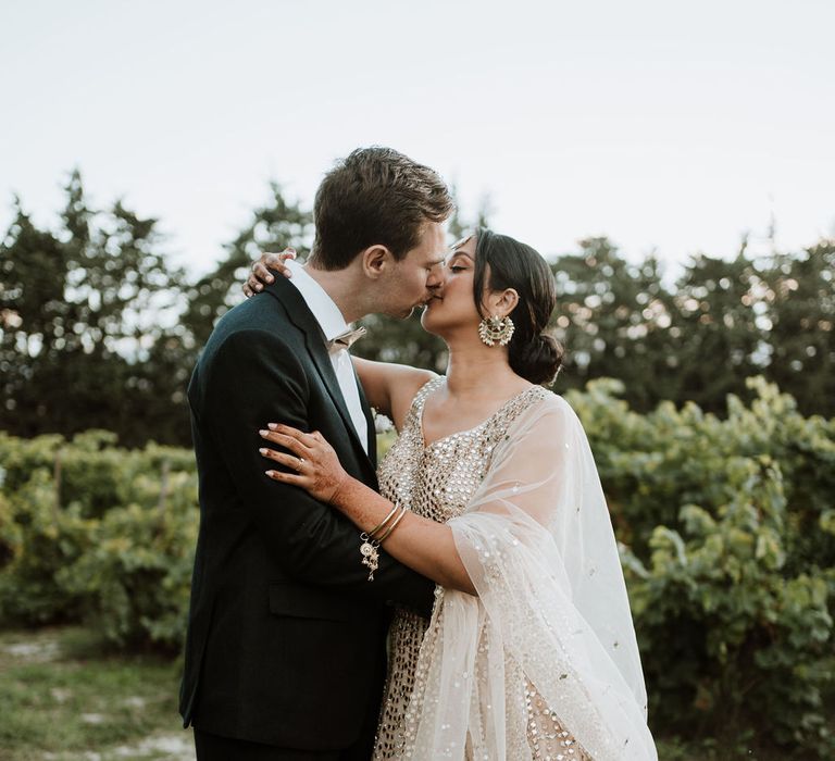 Groom in a black tuxedo kissing his bride in a sparkly wedding dress at their Domaine de Blanche Fleur destination wedding 