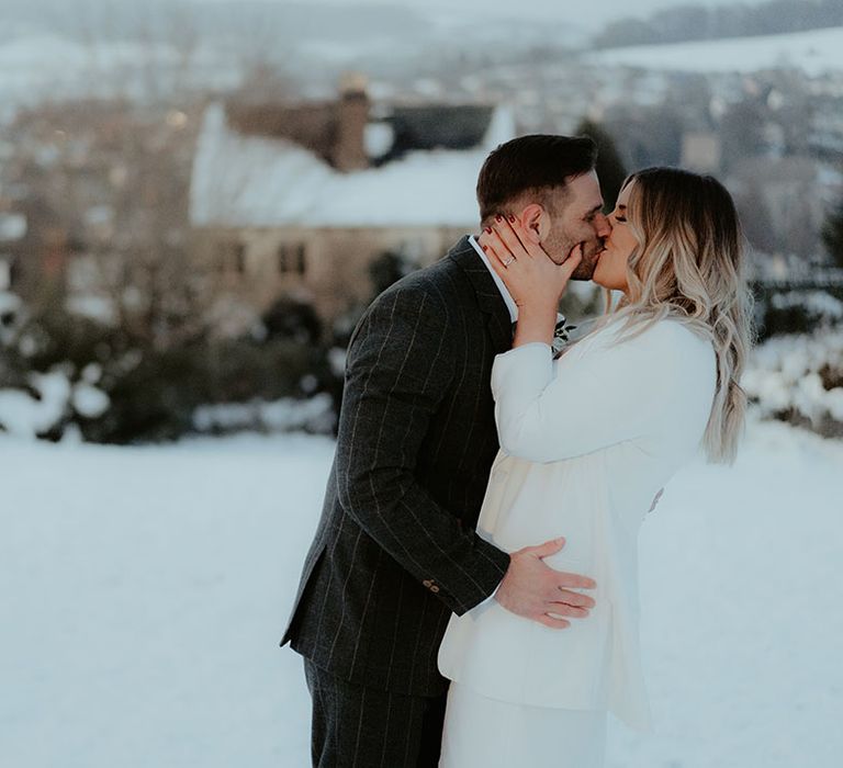 Snowy winter wedding at Christmas with the bride in a white blazer kissing the groom in a dark grey checkered suit 