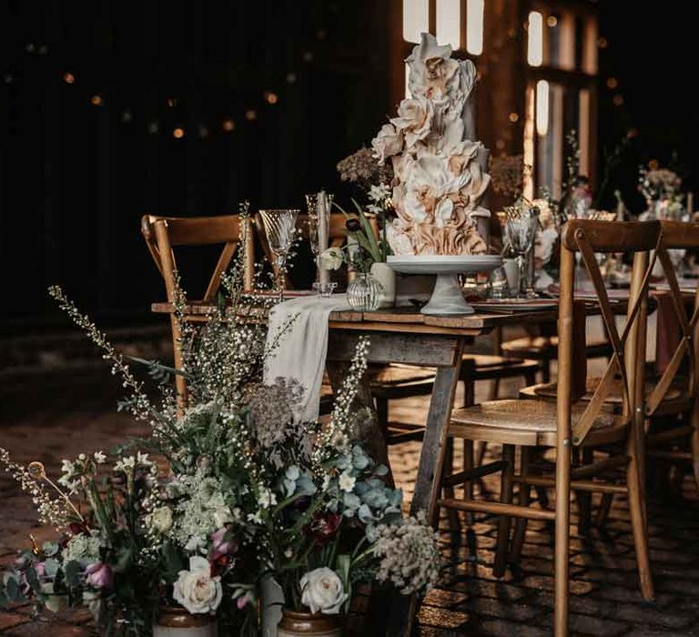 Modern rustic luxe wedding tablescape with white fabric table runner, large foliage, baby's-breath, garden rose and eucalyptus floral arrangements, a large artistic layered wedding cake, tapered candles and ceramic vases 