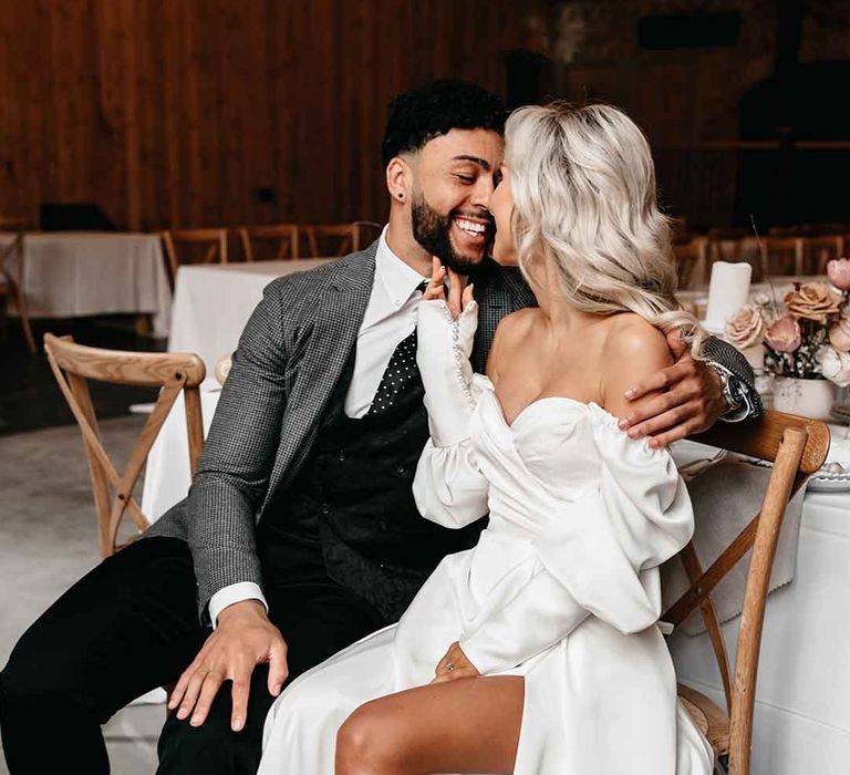 Bride in off the shoulder long sleeve wedding dress with slight cowl neck and front leg slit embracing groom in grey blazer, black waistcoat and navy polkadot tie groom accessory embracing at Lodge Farm wedding venue 