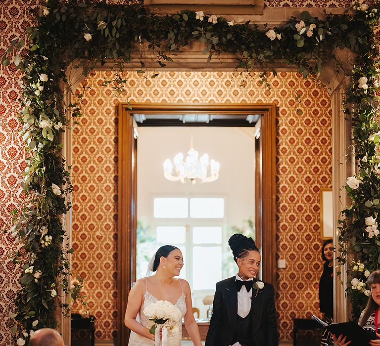 Bride in lace sleeveless v neck Enzoani wedding dress with tulle finish and church length veil and bride in black snakeskin print tuxedo with black bowtie and white rose boutonniere doing wedding exit through Grittleton House with white rose and foliage flower arch around the door 