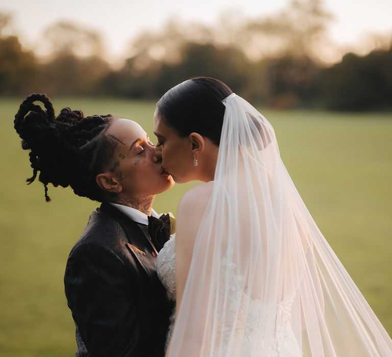 Bride in black snakeskin print tuxedo with black bowtie kissing bride in lace sleeveless v neck Enzoani wedding dress with tulle finish and church length veil on the grounds of Grittleton House wedding venue 