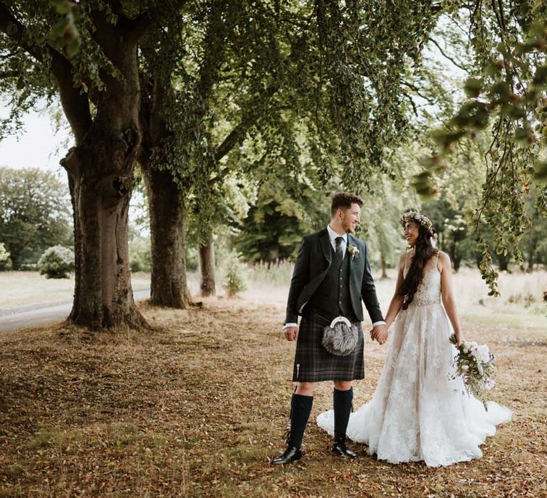 Bride in celestial 3d appliqué sheer overlay wedding dress and bridal flower crown holding large white rose, peony, dried flower and foliage bridal bouquet holding hands with groom in dark grey blazer and waistcoat, dark tartan tie with white flower boutonniere, dark grey and blue tartan kilt and wedding sporran 