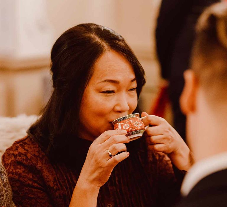 Wedding guest in maroon scallop laced dress sipping tea from tea cup at The Orangery Maidstone wedding venue 