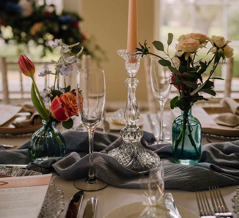 Clear candlestick holder with pastel tapered candle, bright blue glass bottles with dried flowers and champagne glasses on classic white wedding tablescape 