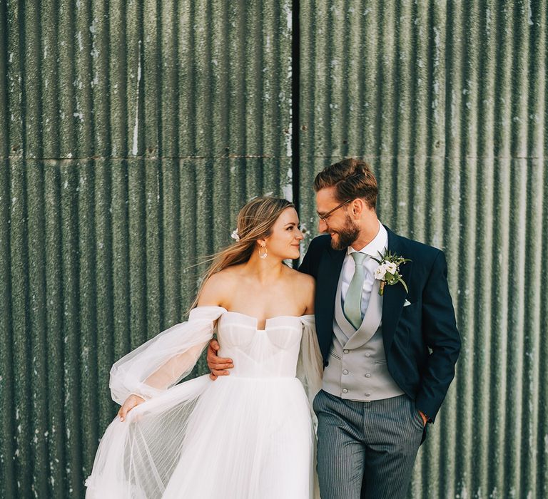 Bride in whimsical romantic long sleeve off the shoulder tulle wedding dress by Alena Leena gazing into the groom's eyes in a morning suit 