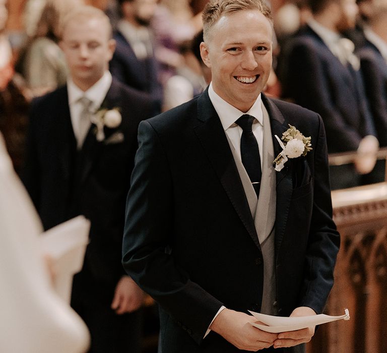 Smiling groom in a three piece wedding suit with a white flower buttonhole holding his wedding order of service booklet 