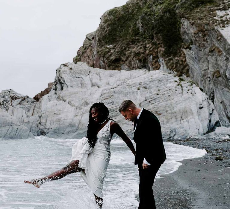 Bride holding up Pronovias lace wedding dress playing in the sea with groom in velvet blazer 