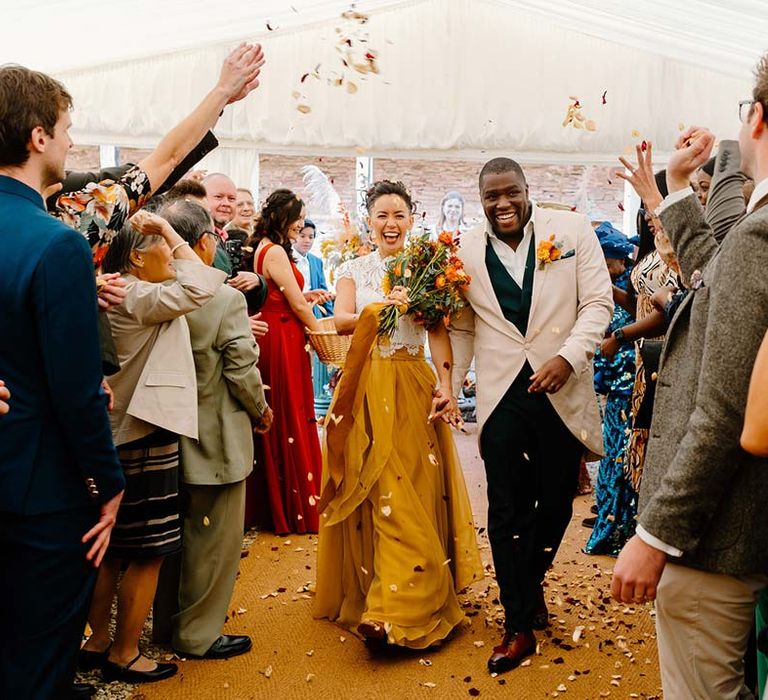 Bride in white mesh lace bridal crop top and yellow wedding skirt walking with groom in cream morning suit with forest green grooms waistcoat and trousers, forest green pocket square and orange boutonniere at Broadfield Court wedding doing colourful dried petal confetti exit