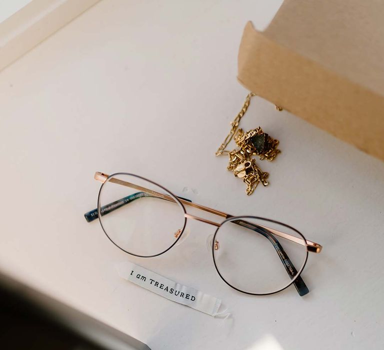 Gold-rimmed grooms glasses with gold bridal jewellery and wedding affirmations printed on paper at Broadfield Court wedding venue