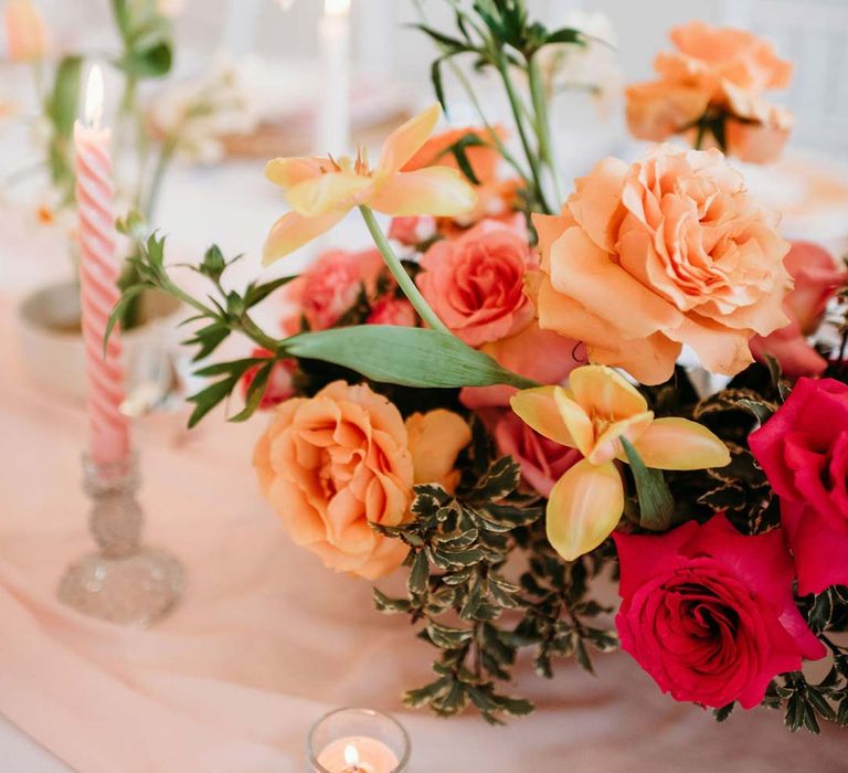 Wedding table with pastel pink twisted tapered candles, tea light candles and flower arrangement centrepiece with pink and orange garden roses, orange floribunda, yellow ranunculus flowers, foliage and Iceland poppies 