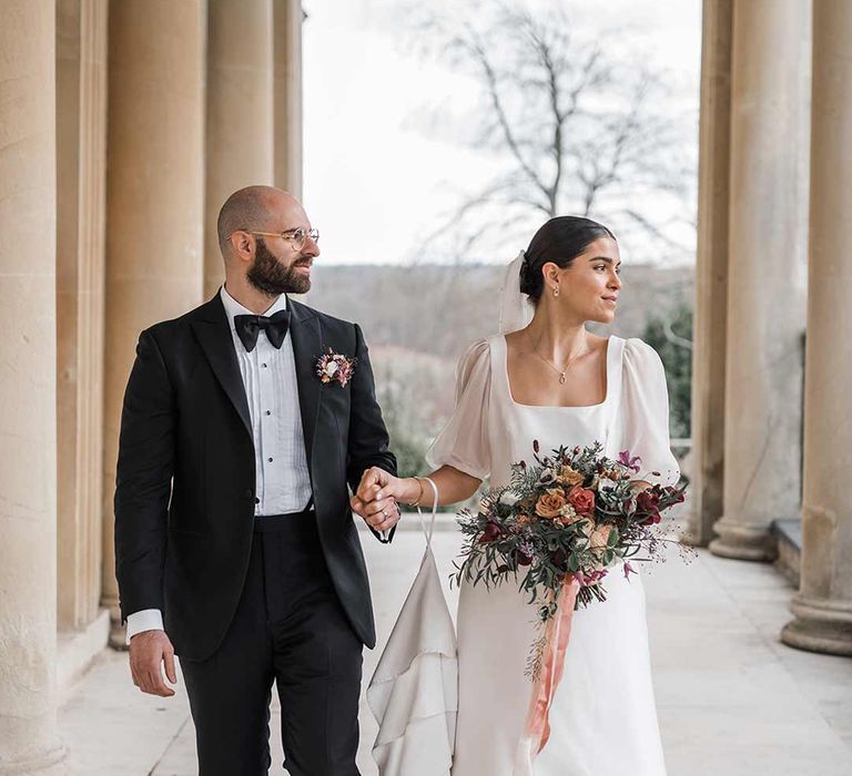 Bride in short sheer puff sleeve wedding dress with square neck and wrist loop holding dried wildflower bouquet walking with groom in classic black tuxedo with bowtie and wildflower boutonniere walking hand in hand at Buxted Park wedding venue