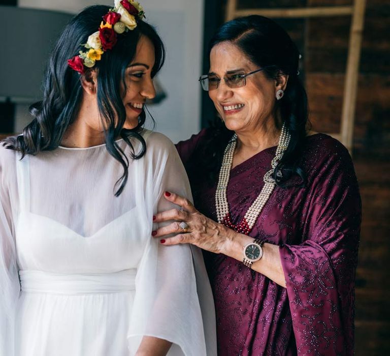 Bride in long sleeve mesh overlay wedding dress and colourful bridal flower crown standing with mother of the bride in deep purple velvet outfit 